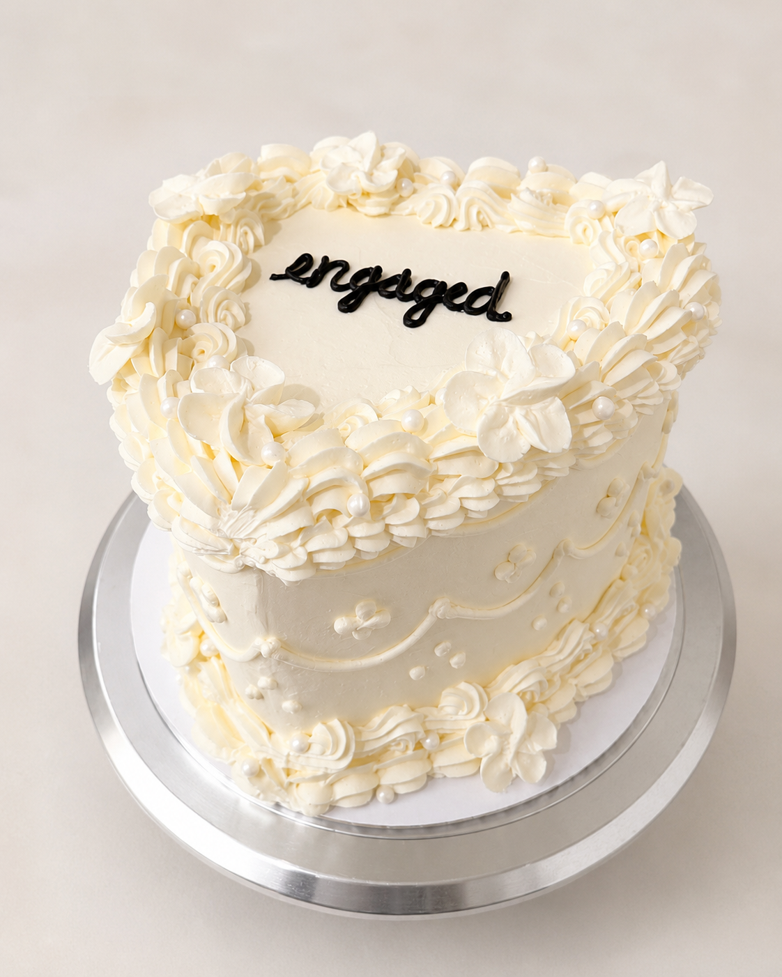 Ivory heart-shaped engagement cake with piped florals, pearl details, and hand-lettered engaged text